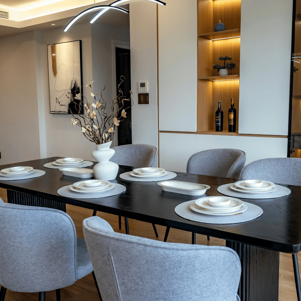 A modern dining room with a dark wooden table set for six, featuring white plates and placemats. Light gray chairs surround the table, and a white vase with branches serves as the centerpiece. Shelves with decor and wine bottles are in the background.