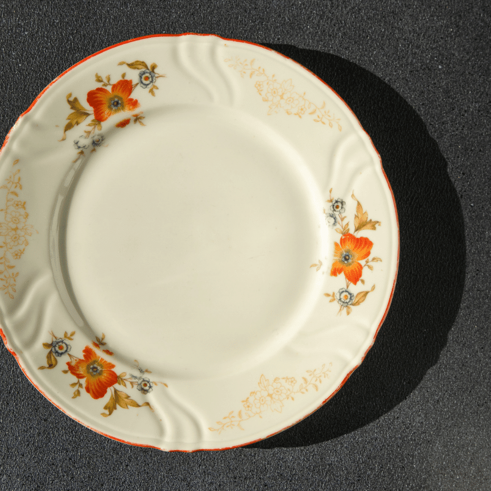 A cream-colored ceramic plate with an orange floral pattern and gold detailing on the rim, displayed on a dark textured surface with a shadow to the right.
