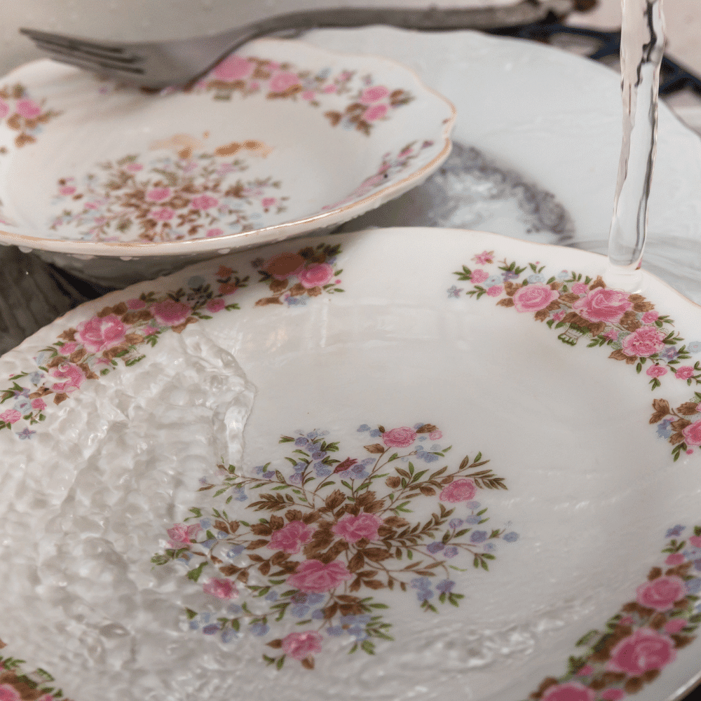 Two floral-patterned porcelain plates are being washed under running water, with a fork resting on the rim of the top plate. The plates feature pink, purple, and blue flowers with green leaves.
