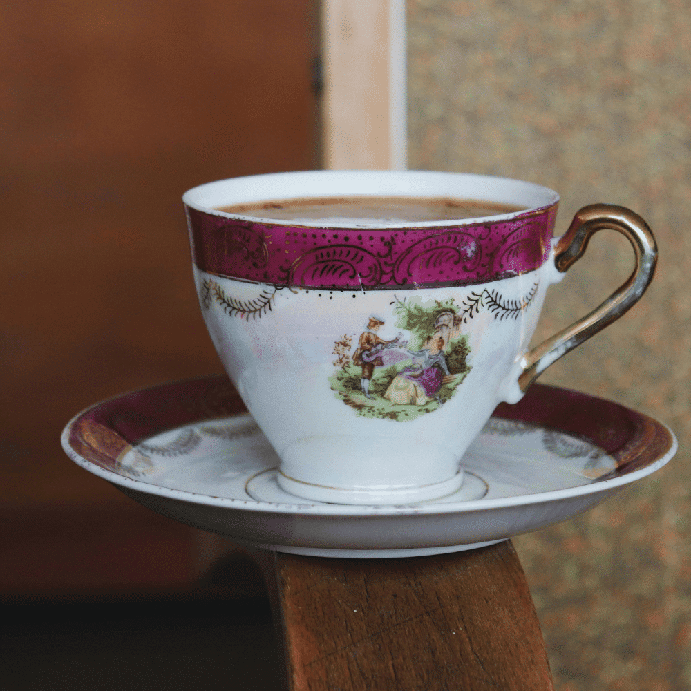 A decorative teacup filled with coffee sits on a matching saucer. The cup features an ornate pattern with gold accents and a scene of people in vintage attire. The set rests on a wooden surface.