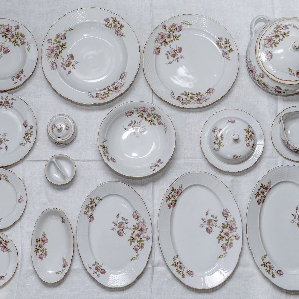 A set of white porcelain dishes with pink floral patterns and gold trim, including plates, bowls, serving dishes, a teapot, and covered containers, all arranged neatly on a white tablecloth.