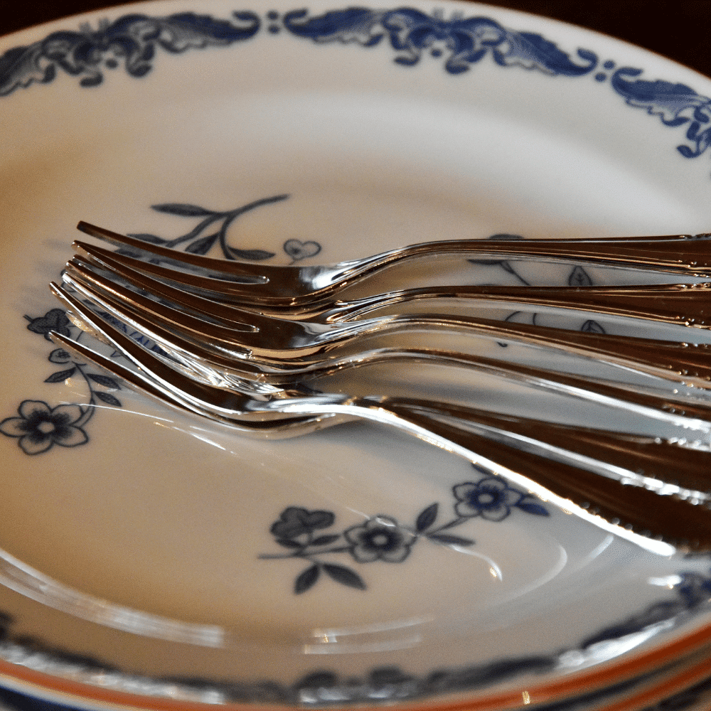 Six silver forks are stacked on top of a white ceramic plate with blue floral patterns along the border and center. The image focuses on the utensils and decorative dishware.
